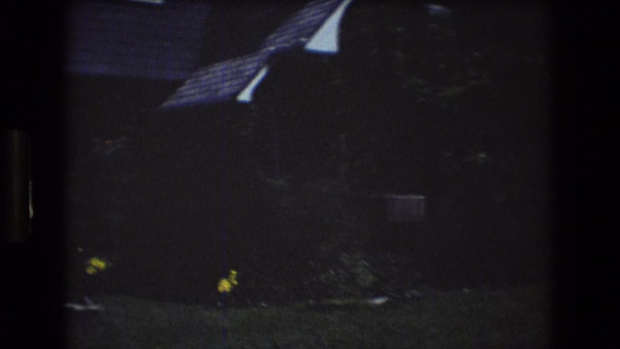 1976 Mt Bromley, Vermont frame showing a dim residential exterior with a sloped roofline, white house edge, shaded lawn, shrubs, and yellow flowers.