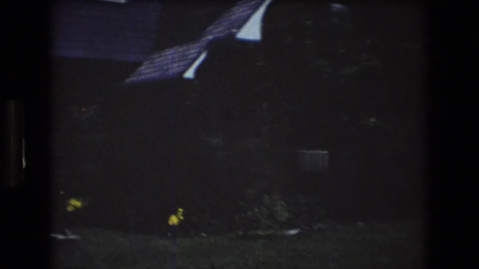1976 Mt Bromley, Vermont frame showing a dim residential exterior with a sloped roofline, white house edge, shaded lawn, shrubs, and yellow flowers.