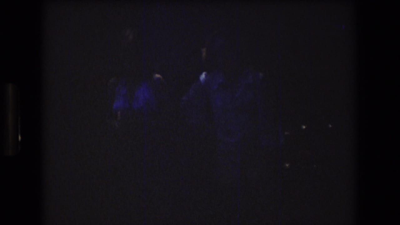 Dark 1976 8mm frame from the supplied Mt Bromley, Vermont clip with faint blue-purple illumination and indistinct adult figures beginning to emerge.