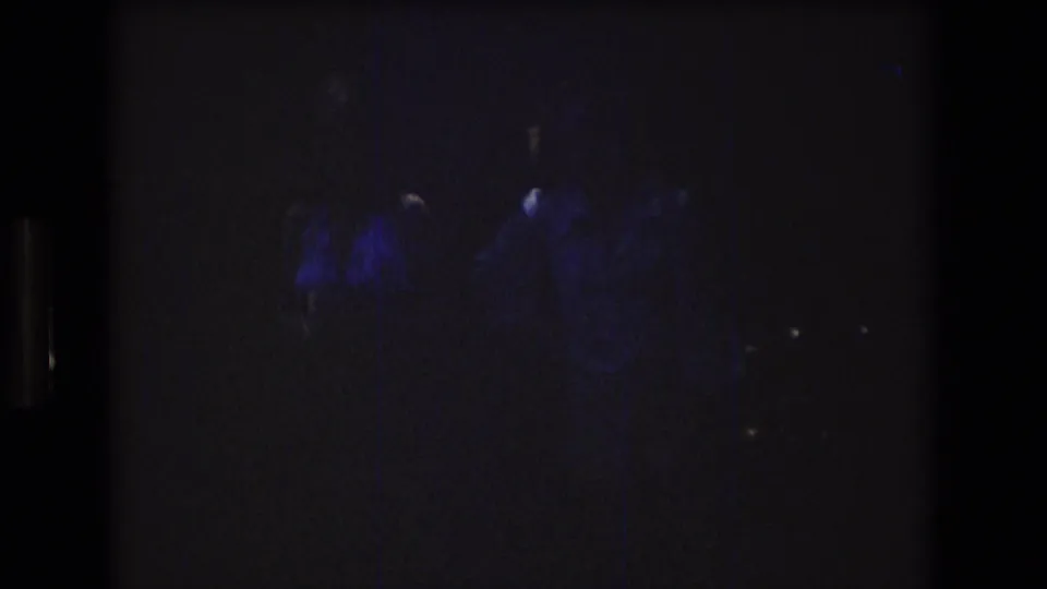 Dark 1976 8mm frame from the supplied Mt Bromley, Vermont clip with faint blue-purple illumination and indistinct adult figures beginning to emerge.