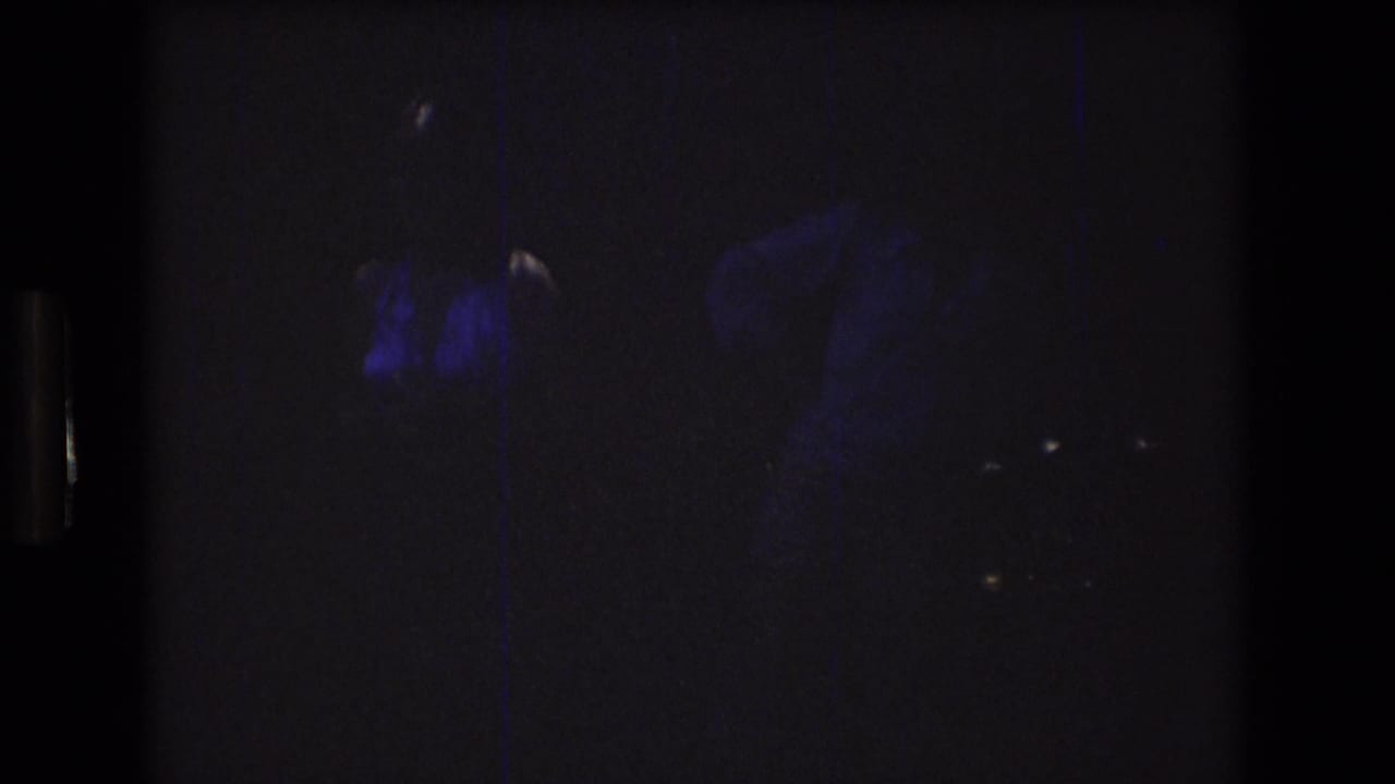 Low-light 8mm frame shows two faint adult figures outdoors at night, with blue-purple exposure and heavy grain obscuring facial detail.
