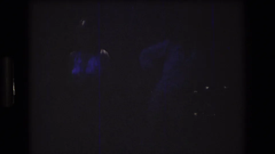Low-light 8mm frame shows two faint adult figures outdoors at night, with blue-purple exposure and heavy grain obscuring facial detail.
