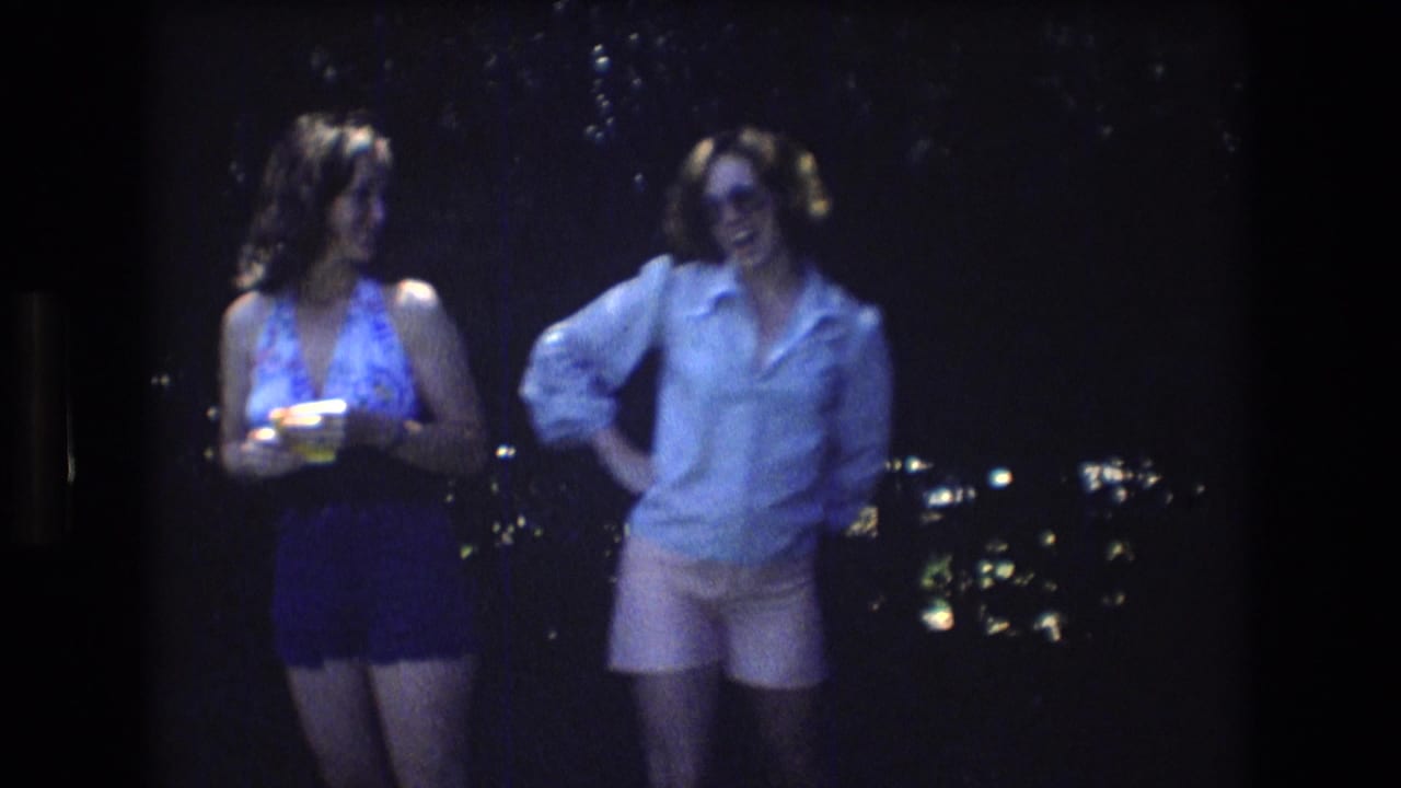 In the supplied 1976 Mt Bromley home-movie frame, two adult women stand outdoors at night, one in a patterned halter top holding a drink and the other in a light shirt, pink shorts, and sunglasses.