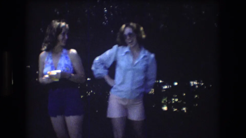 In the supplied 1976 Mt Bromley home-movie frame, two adult women stand outdoors at night, one in a patterned halter top holding a drink and the other in a light shirt, pink shorts, and sunglasses.