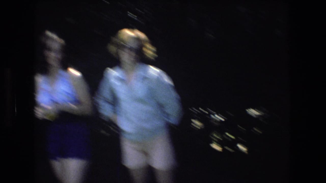 Blurred 8mm frame from the 1976 Mt Bromley clip shows two adult women standing outdoors at night against a dark background with scattered distant lights.