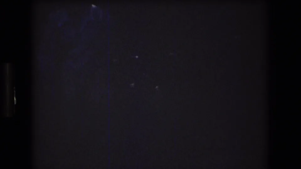 Dark, underexposed 8mm frame from the start of the 1976 Mt Bromley clip, with faint blue-toned shapes and heavy grain but no clear subject visible.