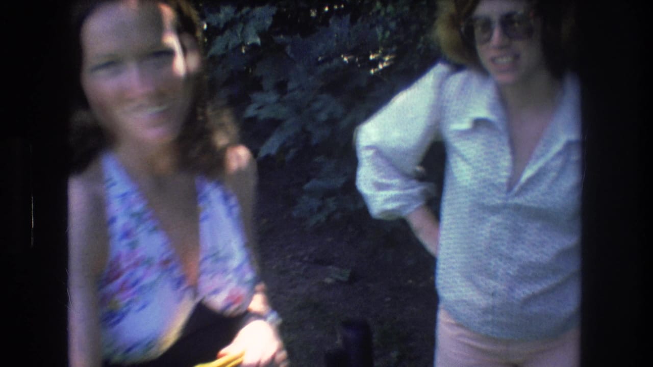 In the 1976 Mt Bromley home-movie frame, one adult woman in a floral top is close to the camera while another woman in sunglasses and a light blouse stands near dense greenery.