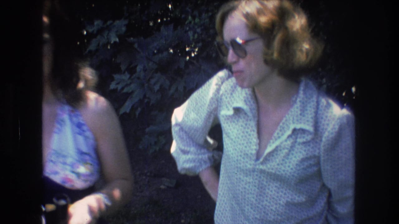 In 1976 Mt Bromley, Vermont 8mm footage, two adult women stand in a wooded area with one woman partly cropped and the other wearing tinted glasses and a light patterned shirt.
