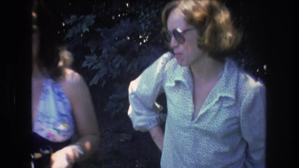 In 1976 Mt Bromley, Vermont 8mm footage, two adult women stand in a wooded area with one woman partly cropped and the other wearing tinted glasses and a light patterned shirt.