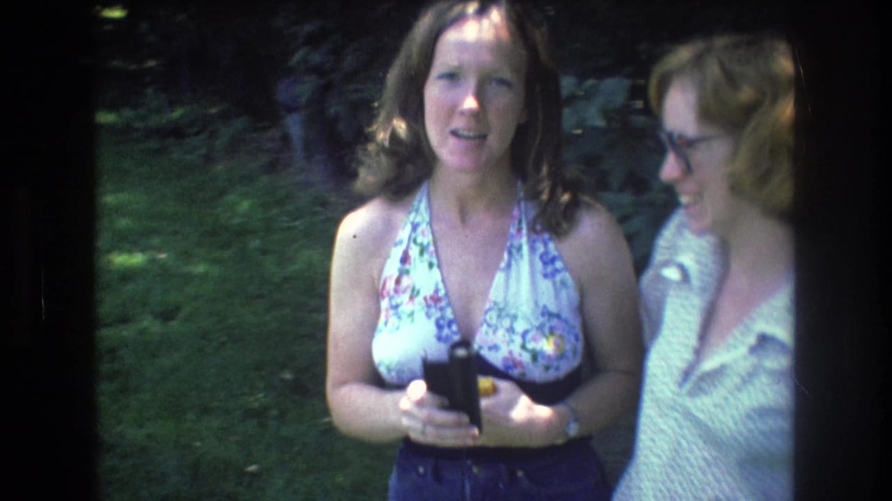 In the Vermont home-movie frame, a woman in a floral halter top faces the camera holding a small black object while a woman in glasses smiles at her side.
