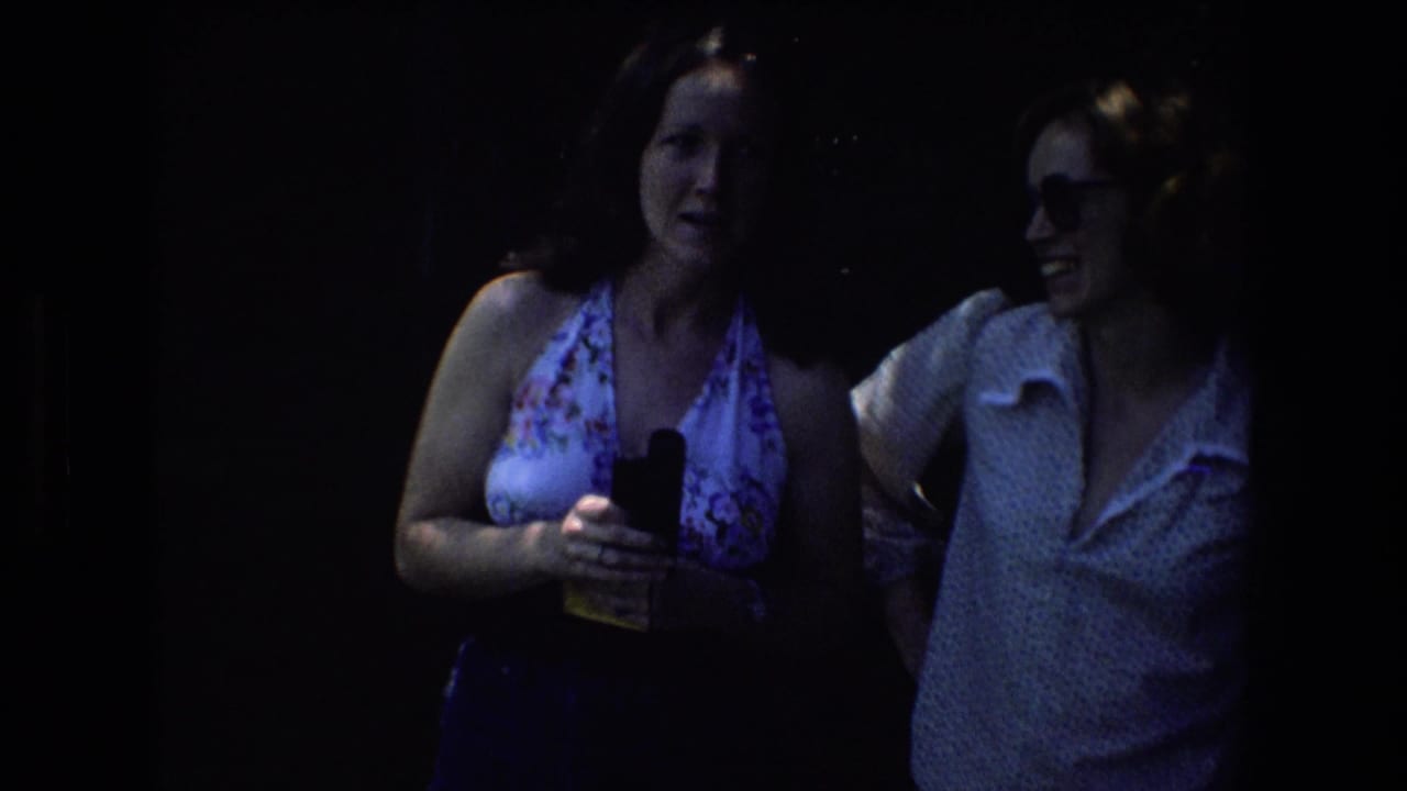The 8mm frame shows the two adult women in heavier wooded shade, with the woman in the floral halter top holding a small black object while the woman in glasses stands nearby.