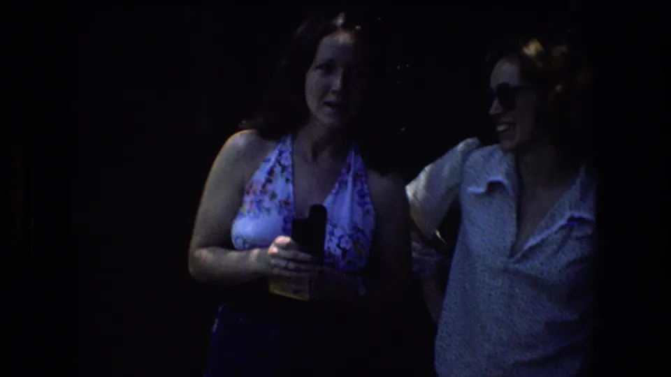 The 8mm frame shows the two adult women in heavier wooded shade, with the woman in the floral halter top holding a small black object while the woman in glasses stands nearby.