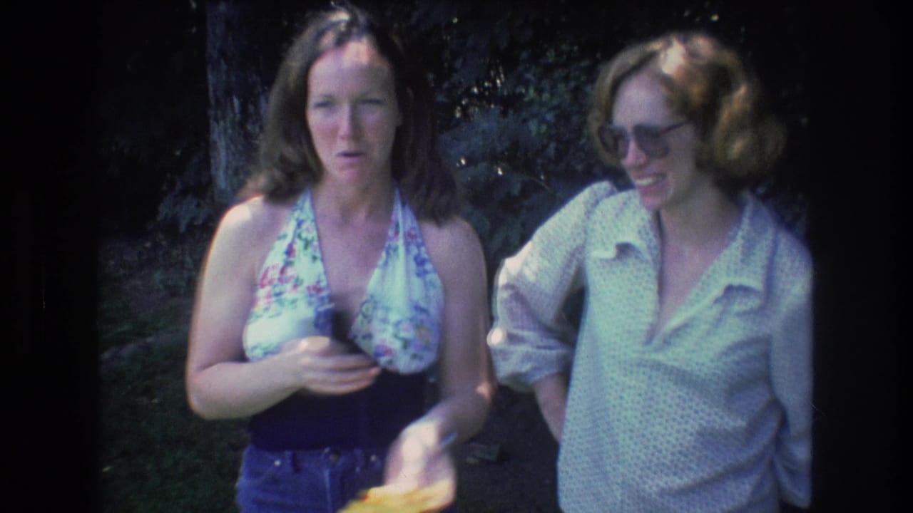 At the end of the 1976 Mt Bromley clip, the woman in the floral halter top gestures while holding a small yellow item as the woman in glasses smiles beside her in the wooded area.
