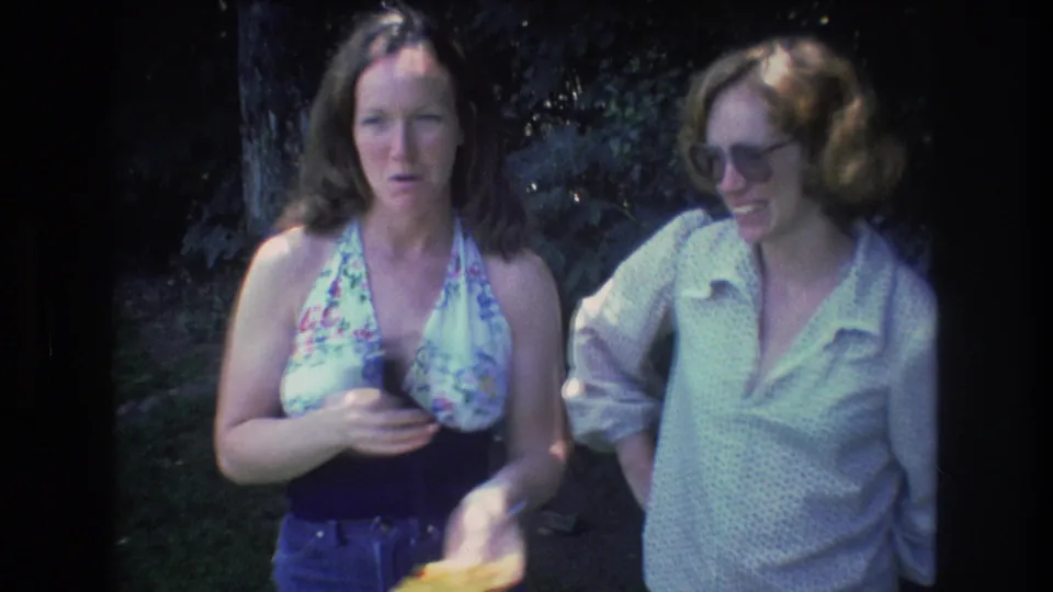 At the end of the 1976 Mt Bromley clip, the woman in the floral halter top gestures while holding a small yellow item as the woman in glasses smiles beside her in the wooded area.
