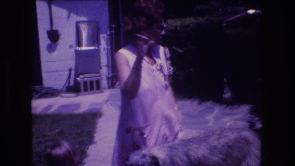 The woman appears to gesture with one hand, likely holding or smoking a cigarette, while the shaggy dog remains nearby in the 1976 Vermont backyard home-movie scene.