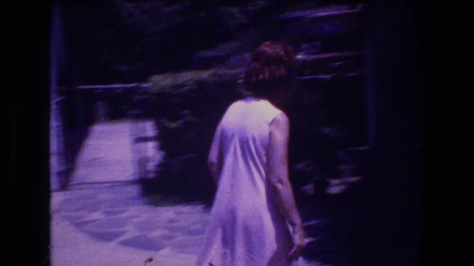 The woman is seen from behind in her light dress as she walks away along the backyard path, with lawn, shrubs, and dark film borders visible in the 1976 Mt Bromley footage.