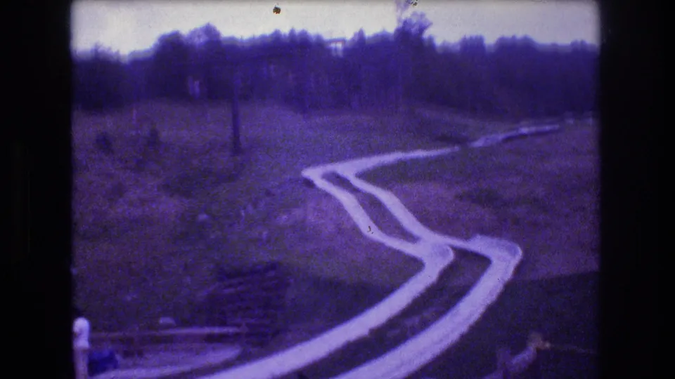 1976 Mt Bromley, Vermont home-movie frame showing a winding concrete slide running down a grassy hillside below a wooded tree line.