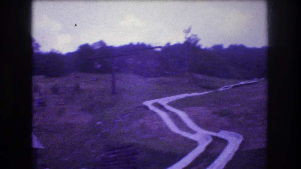 The Mt Bromley hillside slide curves through the frame, with the forest line and small recreation structures visible in soft, purple-tinted 8mm footage.