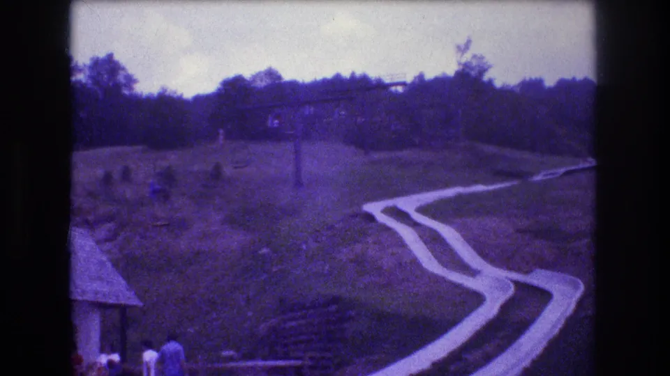Wide 1976 Mt Bromley view of the winding concrete slide, a small building at the base, and several distant visitors on the grassy hillside.