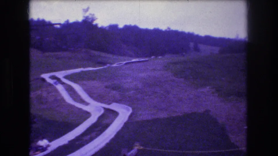 At Mt Bromley, Vermont in 1976, a rider in a small cart appears near the lower part of the winding hillside slide, with open grass and trees beyond.