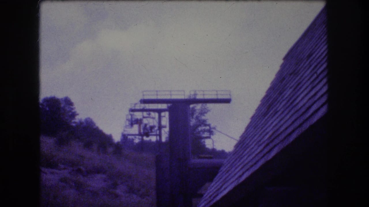 1976 Mt Bromley 8mm frame showing a ski lift tower, chairlift cables, wooded slope, and steep shingled roof beneath an overcast sky.