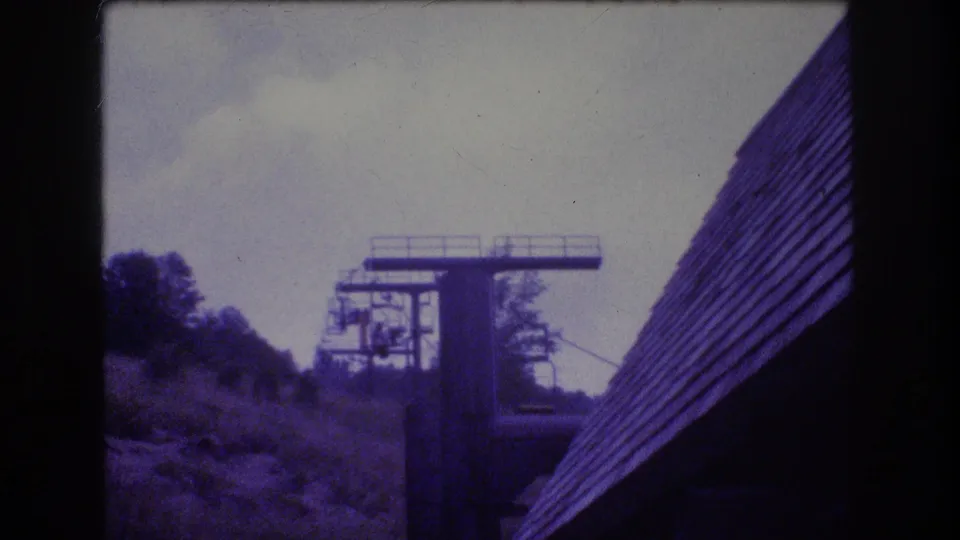 1976 Mt Bromley 8mm frame showing a ski lift tower, chairlift cables, wooded slope, and steep shingled roof beneath an overcast sky.
