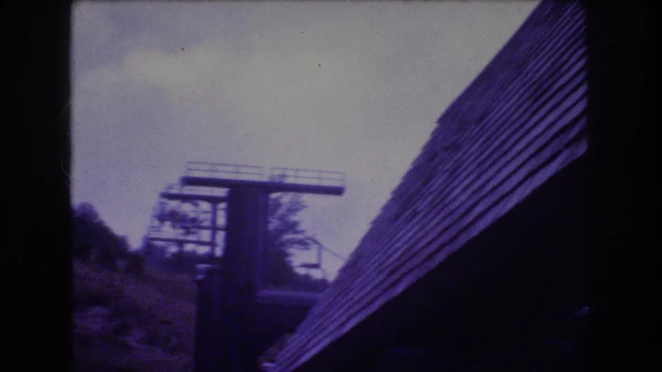 1976 Mt Bromley 8mm frame showing a lift tower partly framed by a steep shingled roof, wooded slope, and pale cloudy sky.