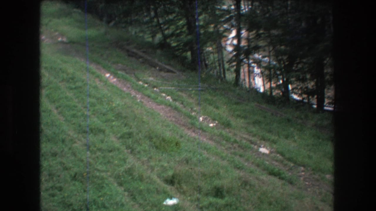 1976 8mm home-movie frame from Mt Bromley, Vermont, showing a green sloped hillside crossed by a narrow dirt track beside thin coniferous trees.