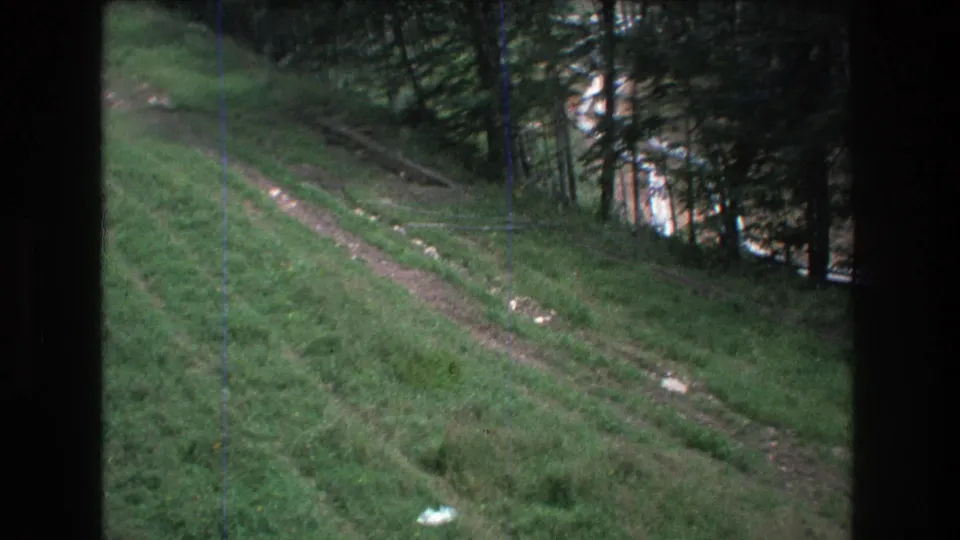 1976 8mm home-movie frame from Mt Bromley, Vermont, showing a green sloped hillside crossed by a narrow dirt track beside thin coniferous trees.