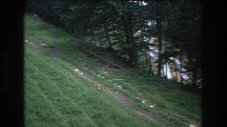 Soft 8mm frame from 1976 Mt Bromley, Vermont, showing a grassy hillside, a curving dirt path, scattered stones, and conifers at the slope edge.