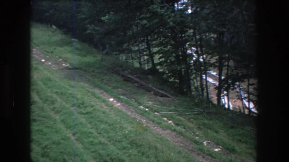 1976 8mm home-movie frame at Mt Bromley, Vermont, with a narrow hillside path, thin coniferous trees, and a pale road or clearing partly visible beyond the trees.