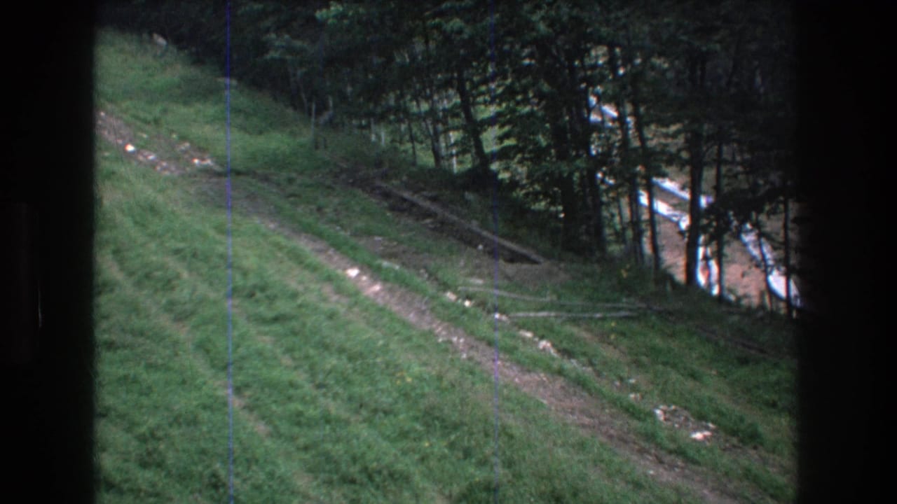 1976 Mt Bromley home-movie frame showing a grassy slope, dirt track, and wooded edge from a moving lift viewpoint.