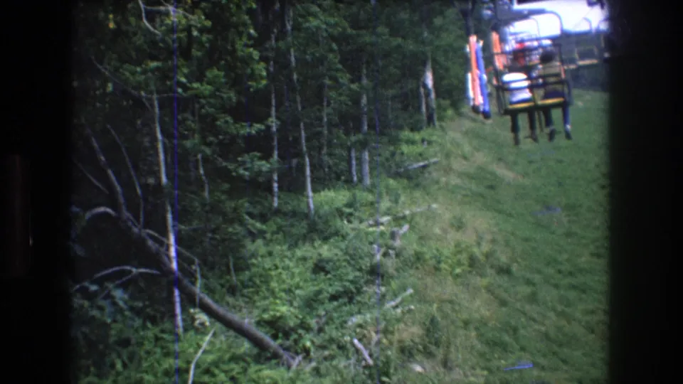 A chair or cable car carrying distant riders appears over the wooded summer slope at Mt Bromley, Vermont.
