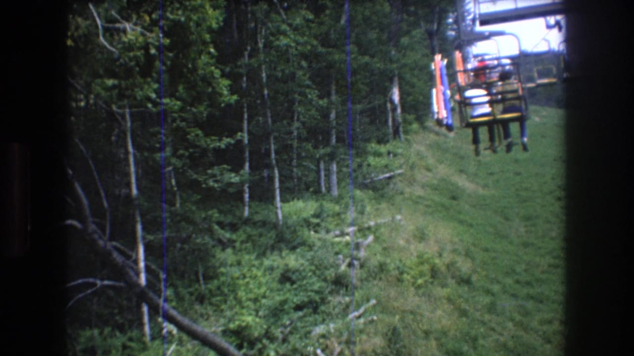 1976 home-movie view at Mt Bromley, Vermont, showing chairlift seats moving up a grassy mountain corridor bordered by dense green forest.
