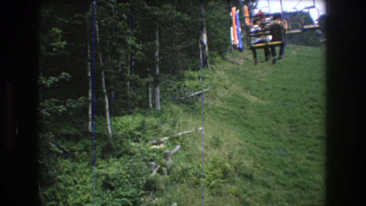 1976 8mm home-movie frame from Mt. Bromley, Vermont, looking from a chairlift toward empty lift chairs, grass, and dense summer trees.
