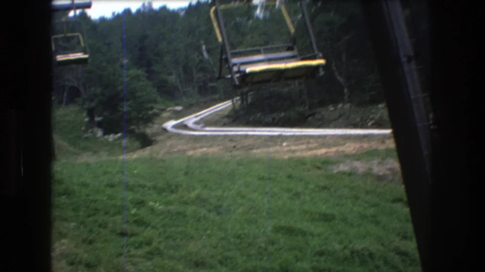A 1976 Mt. Bromley chairlift frame looks down toward a pale winding road or track curving beside trees and a lift support structure.
