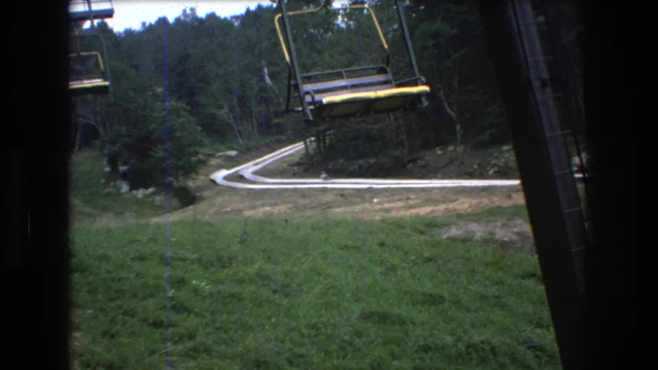 From the chairlift at Mt. Bromley, the frame shows a curving road, forest, lift structure, and a distant person in dark clothing near the lower slope.