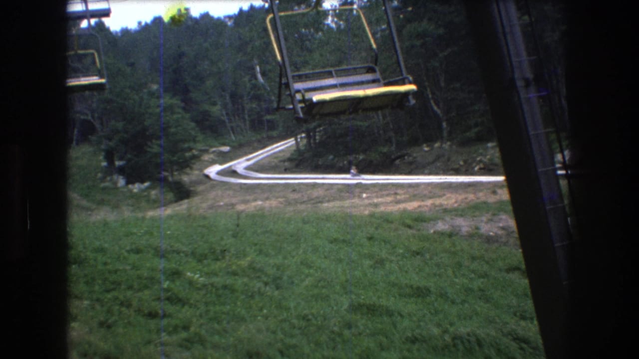 1976 8mm view at Mt. Bromley, Vermont, showing empty open chairlift seats above a winding pale road bordered by grass and dense trees.
