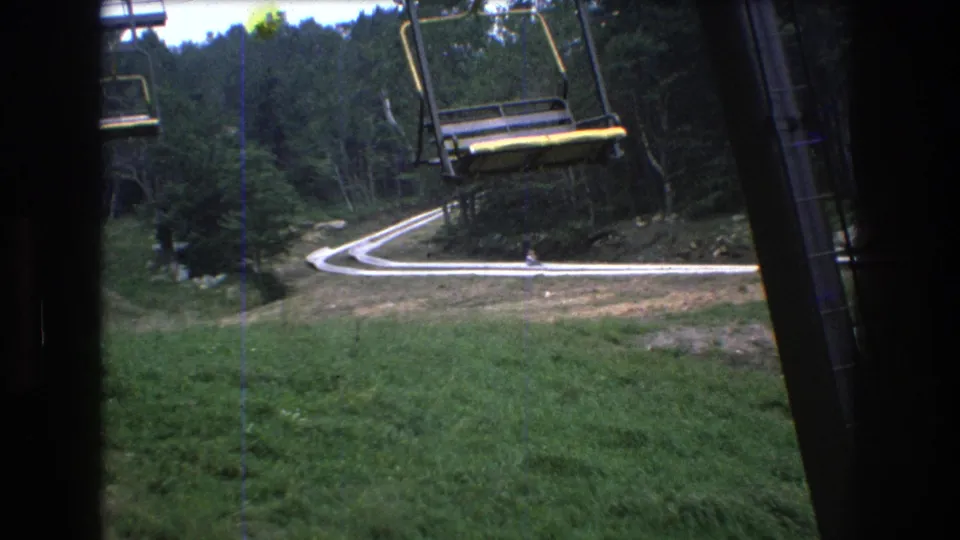 1976 8mm view at Mt. Bromley, Vermont, showing empty open chairlift seats above a winding pale road bordered by grass and dense trees.