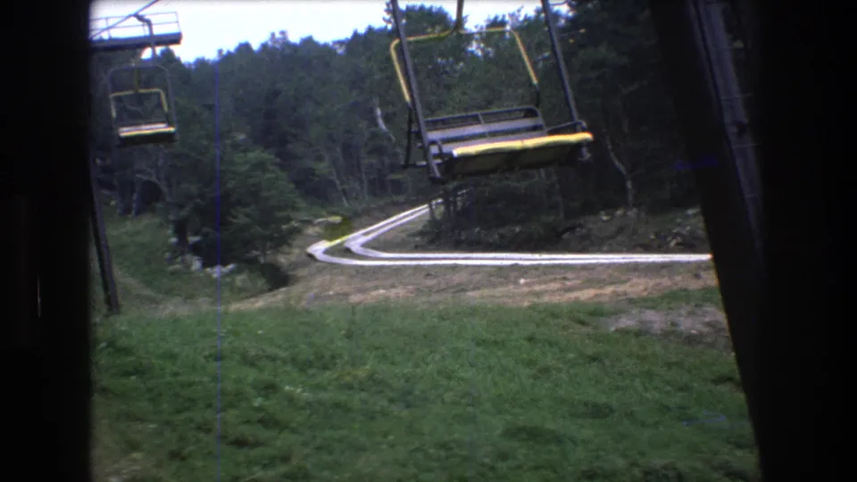 A low open chairlift seat dominates the 8mm frame as the lift passes over grass and a winding road at Mt. Bromley, Vermont.
