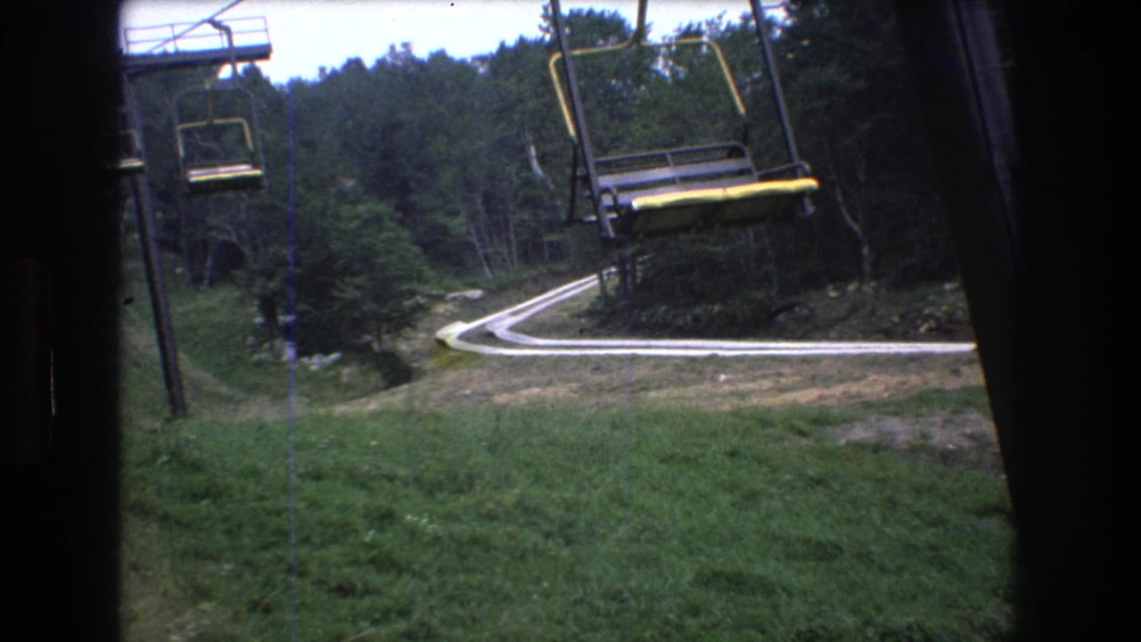 1976 8mm home-movie frame from Mt. Bromley, Vermont, showing yellow-and-black chairlift seats over a green slope with trees and a curved path below.