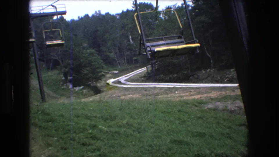 1976 8mm home-movie frame from Mt. Bromley, Vermont, showing yellow-and-black chairlift seats over a green slope with trees and a curved path below.