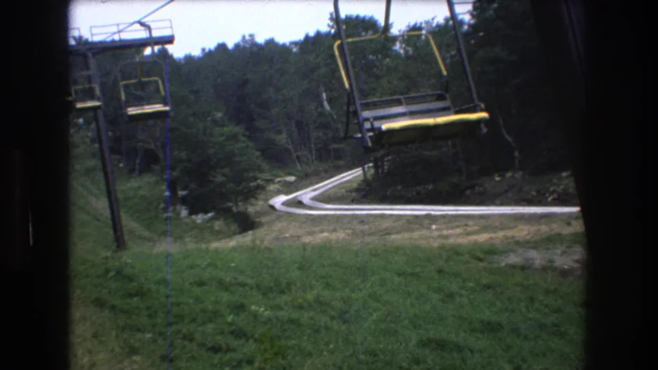 1976 8mm frame at Mt. Bromley showing suspended chairlift seats and cables above a grassy mountain slope with a curving path and wooded background.