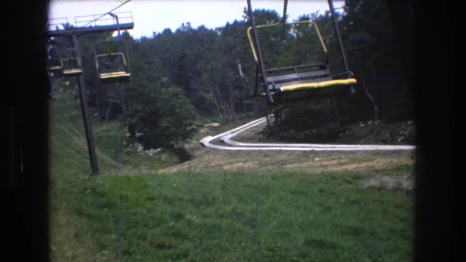 1976 8mm home-movie view from a moving chairlift at Mt. Bromley, with lift chairs, a green slope, forest, and a winding pale path visible below.