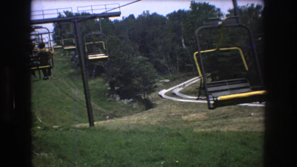 1976 Mt. Bromley 8mm frame showing a chairlift seat close to the camera, additional chairs along the cable, and a green resort slope with trees and a winding path below.