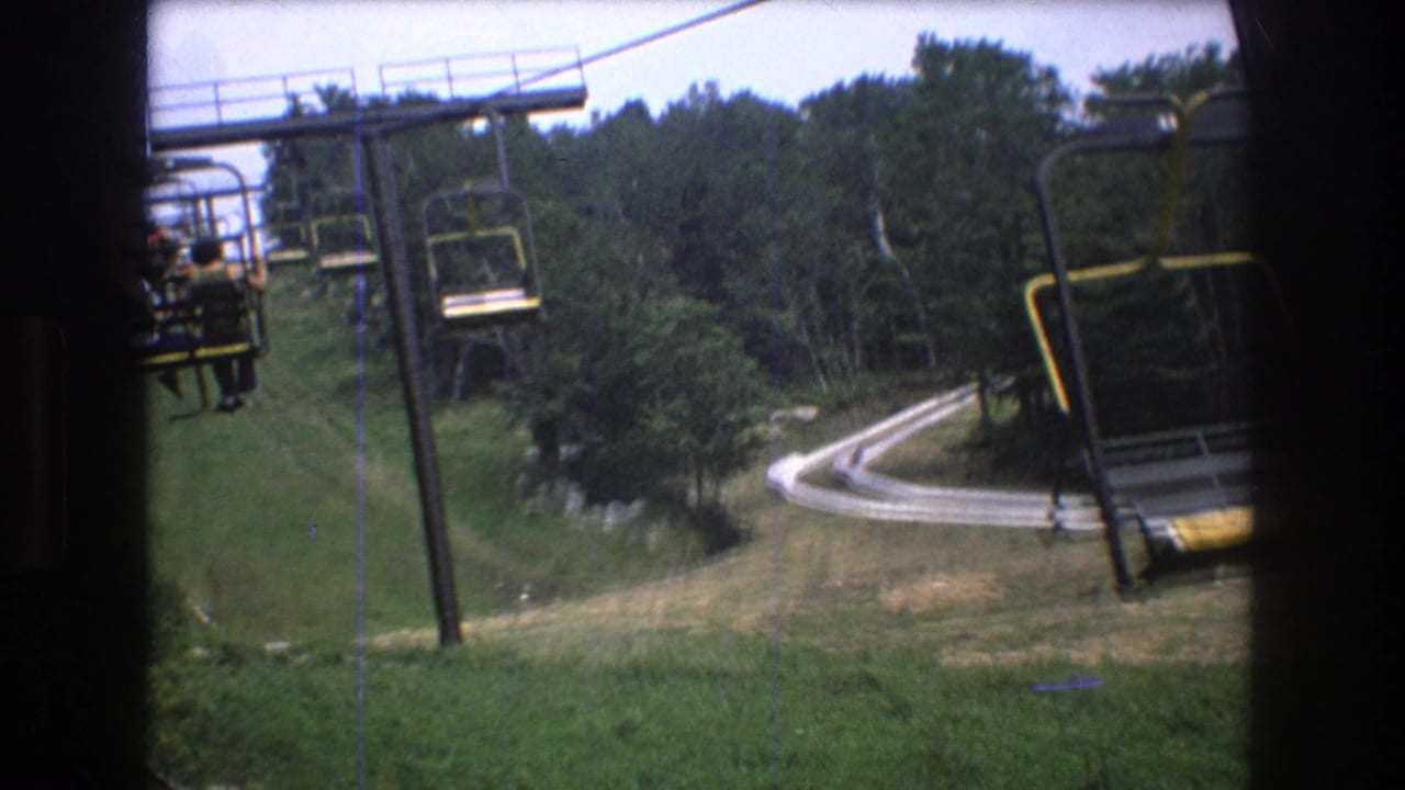 1976 Mt Bromley 8mm frame from a moving chairlift, showing yellow-and-black lift chairs above a green slope, dense trees, and a winding pale track or road below.