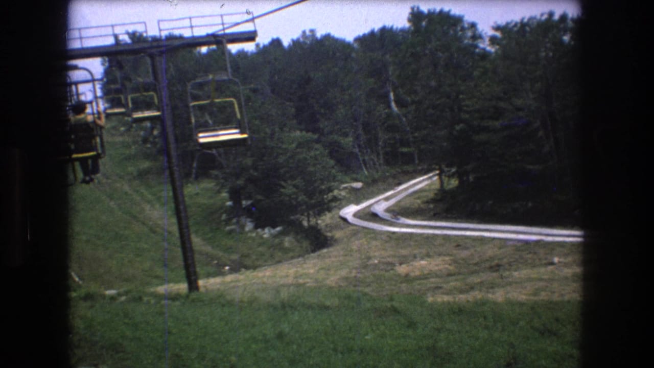 Open chairlift seats hang above a wooded hillside with a curving pale track below.