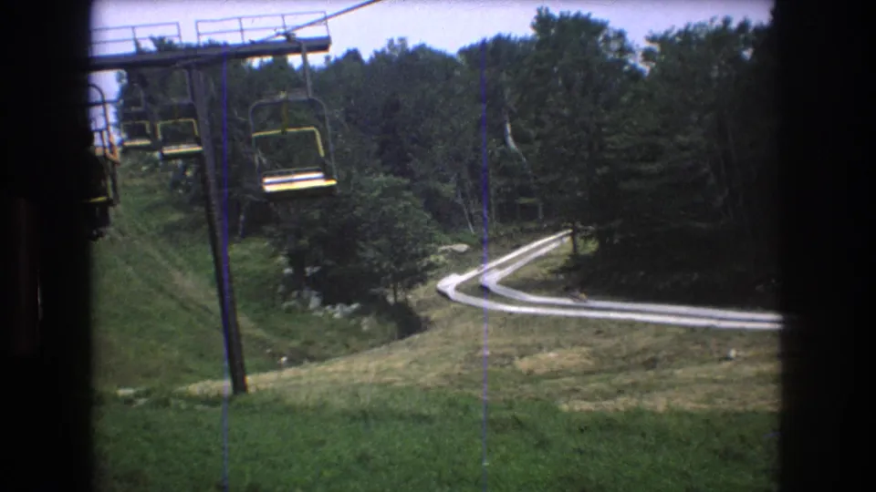 8mm home-movie frame from the Mt Bromley chairlift, showing a grassy ski-area slope, lift tower, empty suspended chairs, trees, and a winding pale track beyond.
