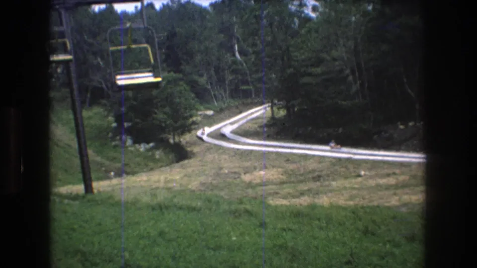 Mt Bromley chairlift view from 1976, with a suspended yellow chair, grassy slope, dense evergreen and deciduous trees, and small distant riders or visitors on the curving track below.