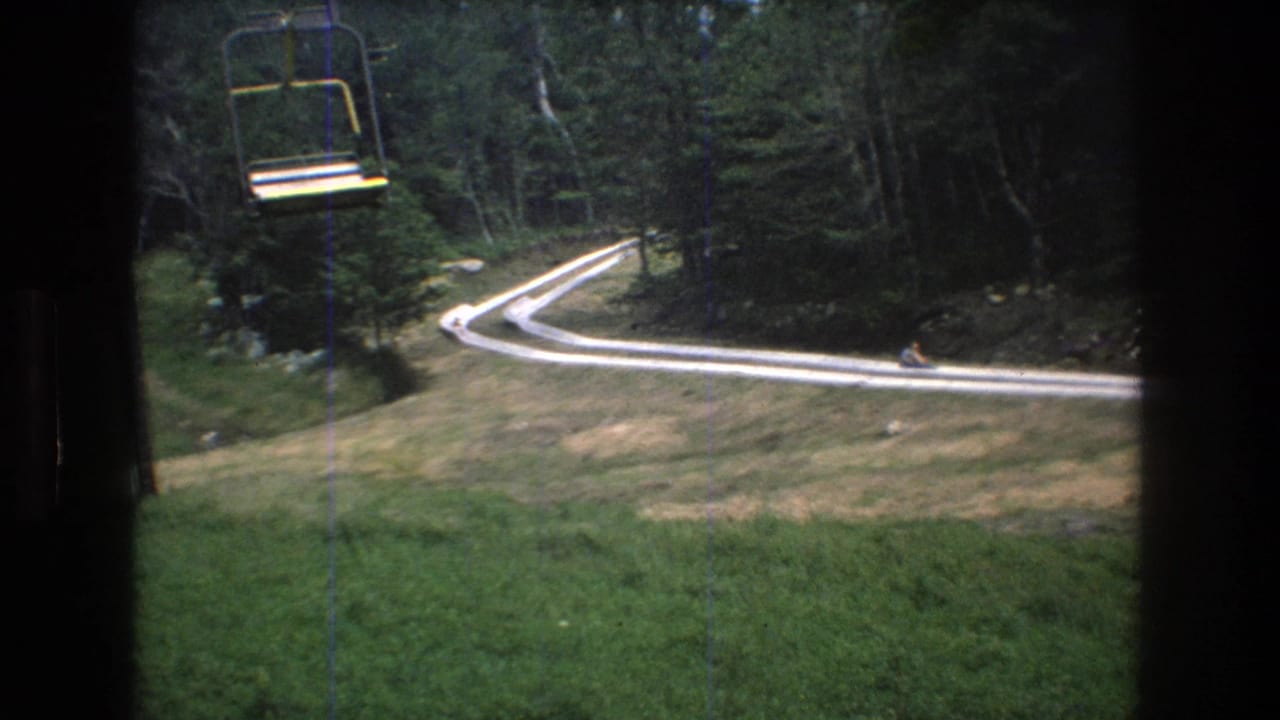 1976 Mt. Bromley home-movie frame showing a yellow chairlift seat passing over a grassy slope with a winding white track bordered by dense trees.